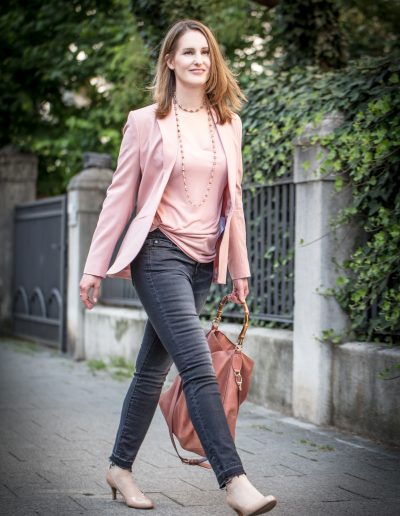 Frau im Business Outfit. Mittelblonde Haare. Graue Jeans, nudefarbener Blazer, nudefarbenes Shirt, rosewoodfarbene Ledertasche mit Bambusgriff. Nudefarbene Pumps. Kette aus Muranoglasperlen.