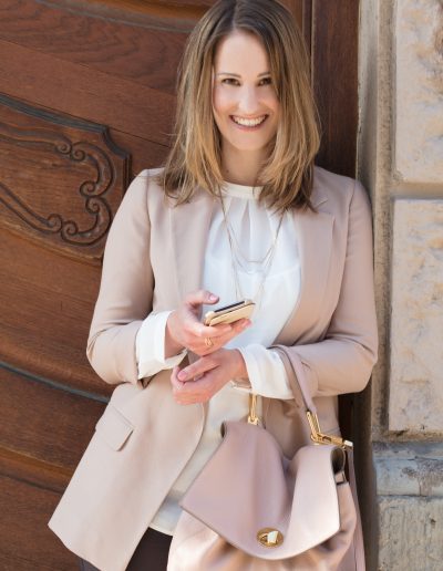 Frau im Business Outfit. nudefarbener Blazer,weiße Seidenbluse, nudefarbene Ledertasche, braune Hose. Goldener Schmuck. Smartphone in der Hand.