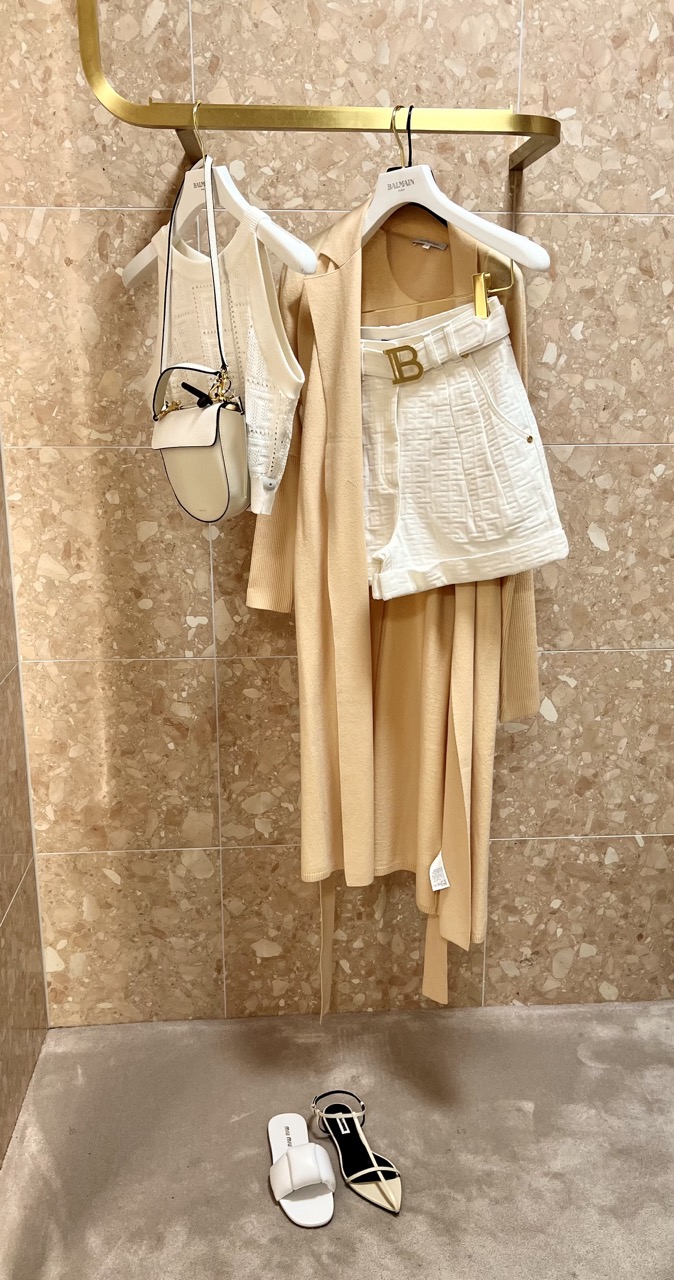 Outfit Kombination auf Kleiderständer mit cremefarbenem Top mit Shorts, Handtasche und Schuhen dazu Vanillefarbener Strickmantel vor beigefarbener Marmor Wand
